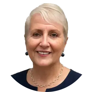 Joane Goulding, founder of the Goulding System with short, white hair, wearing a navy top, gold chain necklace, and black earrings, against a plain light background.